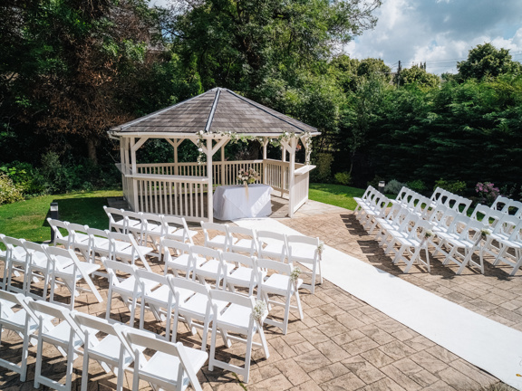 Gazebo in front of lush gardens, rows of white chairs and a white runner carpet face the gazebo.