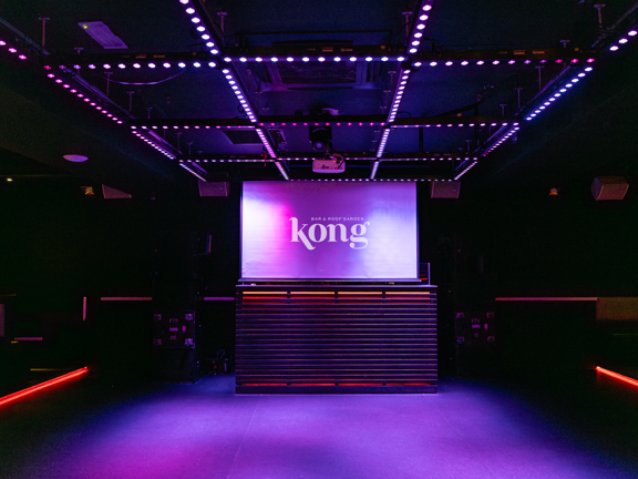 Club room at Kong Glasgow. Black room with stripes of coloured lights across the ceiling and red neon lights around the edge of the floor. A large screen reads Kong.