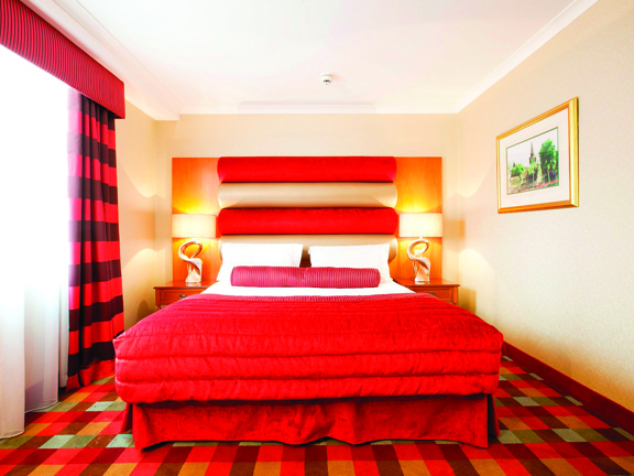 An internal view of a Carlton George bedroom. The room is decorated with a brightly coloured plaid carpet and a red headboard. The bed is decorated with a red cushion and blanket. There is a framed print on the right wall and a window with red curtains on the left.