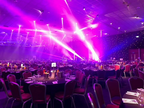 Large dinner set up tables with black tablecloths surrounded by red velvet chairs. Black star cloth behind the stage with purple lights beaming out from the stage and ceiling. 