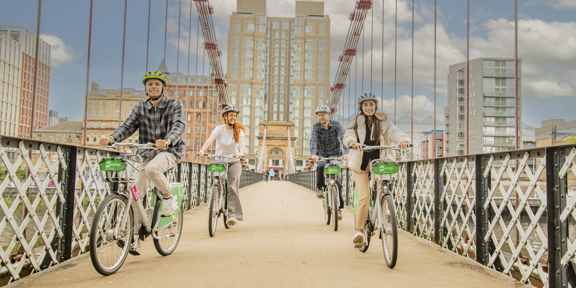 Four cyclists crossing a footbridge with the modern grey building of the Virgin hotel in the background