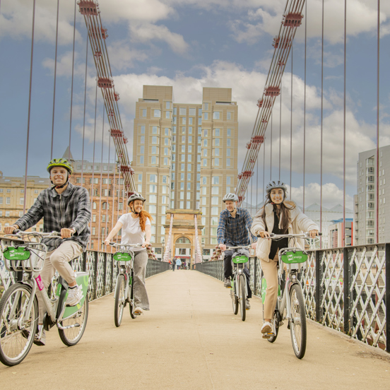 Four cyclists crossing a footbridge with the modern grey building of the Virgin hotel in the background