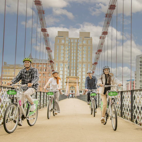 Four cyclists crossing a footbridge with the modern grey building of the Virgin hotel in the background