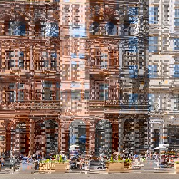 Historic buildings on St Vincent Place in Glasgow, including The Citizen and Anchor Line, with outdoor terrace seating and people dining in front of ornate red sandstone and white façades.