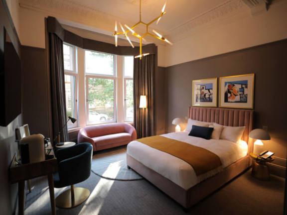 Cosy hotel bedroom with a king-sized bed and a large window. Rich brown walls and curtains with accents of gold and velvet accessories create a warm environment.