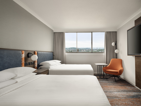 Hotel bedroom with two double beds dressed with crisp white linen and 4 fluffy white pillows. An orange accent chairs sits in teh corner under a large window looking out across the city. 