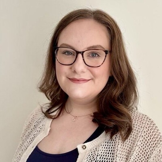 Portrait of a smiling woman with shoulder-length brown hair and glasses, wearing a cream knitted cardigan over a dark top, standing against a plain light-coloured background.