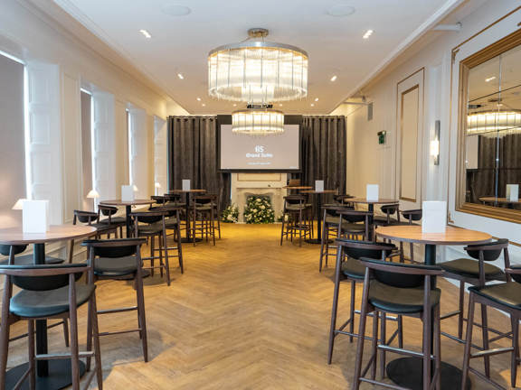 Modern white room set up restaurant style. Chandeliers hang from above and a tv screen at the far end.
