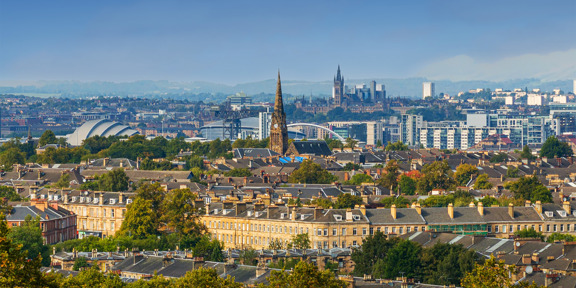 Sunny cityscape with the Gothic revival-style spire of the University of Glasgow's main building, surrounded by greenery and buildings of the West End, including the modern buildings of the SEC Armadillo and the OVO Hydro arena