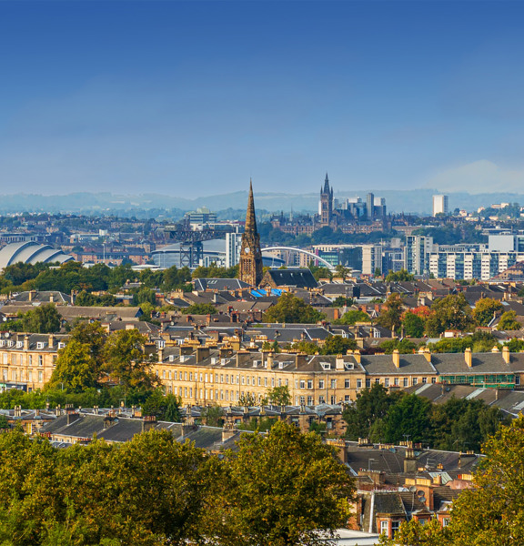 Sunny cityscape with the Gothic revival-style spire of the University of Glasgow's main building, surrounded by greenery and buildings of the West End, including the modern buildings of the SEC Armadillo and the OVO Hydro arena