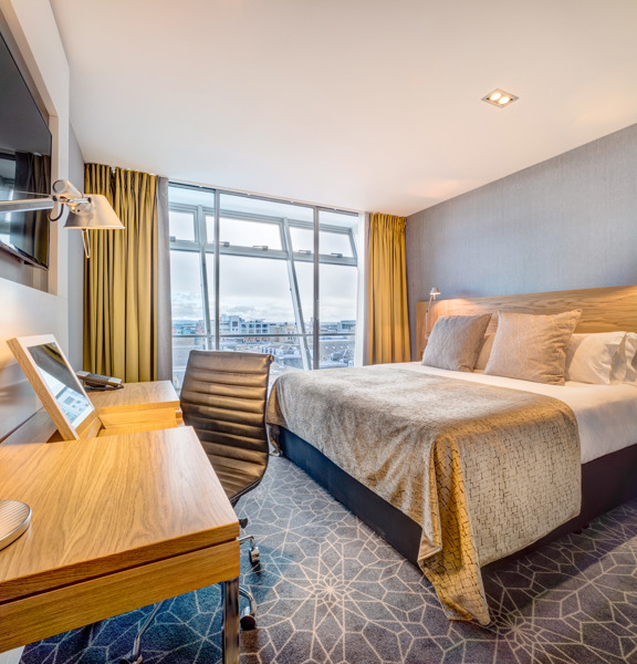 An interior shot of a hotel room, the bed is large and tidy and has a grey blanket and cushion on it. A television, desk and chair are all visible. The far wall is floor to ceiling windows with yellow curtains on either side.