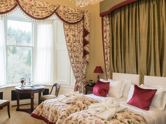 An interior shot of a twin bedroom at Crossbasket Castle hotel. The room has 2 single beds, positioned closely together to the left of the image. The beds and room are decorated opulently with floral throws, curtains and pelmets in cream and magenta. On the end of each bed is a white, plump, folded dressing gown. The bay window has white net curtains and a small round table and 2 wooden chairs sit in the bay. A lit chandelier hangs above the beds and illuminates the plaster ceiling-rose.
