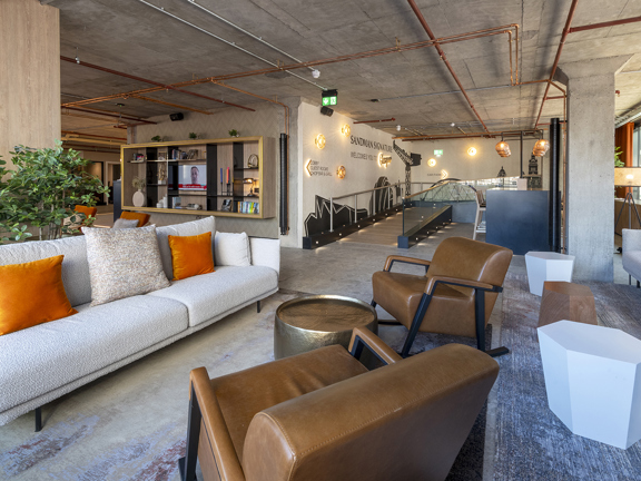 A photo from inside the Sandman hotel lobby. A large open plan space is filled with an eclectic mix of modern sofas, chairs, tables and rugs in shades of grey orange and brown. The walls and ceilings are concrete with pale wood accents and visible copper piping. Floor to ceiling windows with orange velvet curtains can be seen along the right wall and the light in the room suggests the wall behind the photographer is also windowed. A ramp access entrance can be seen ahead, with murals of Glasgow's landmarks.