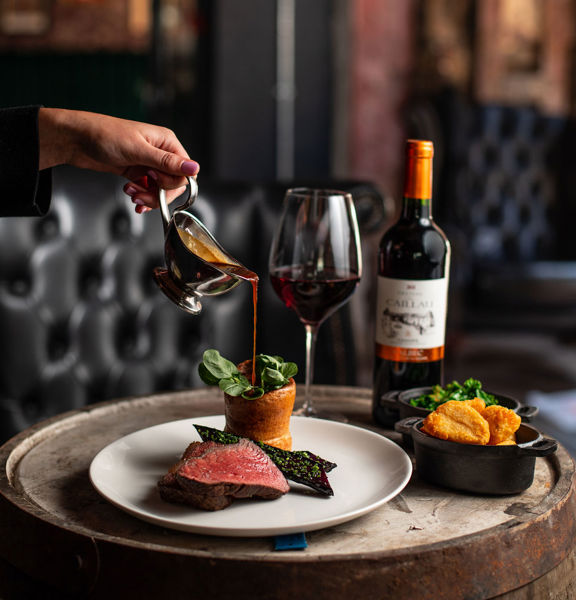 A close up image of a Duke's Umbrella roast dinner placed atop a reclaimed wooden barrel. A white, feminine hand and fore-arm can be seen pouring gravy from a small silver gravyboat onto a clean plate of roasted vegetables, sliced beef and Yorkshire pudding. Rustic metal bowls of roast potatoes and greens, a bottle of red wine and a large wine glass can also be seen on the wooden top. Plush, black leather seating is visible in the background.
