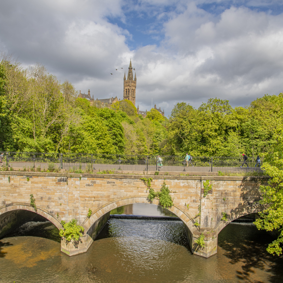 Sunny view of Gothic revival-style spire of the University of Glasgow's main building, surrounded by greenery and with the 3-arched stone Snow Bridge in the foreground