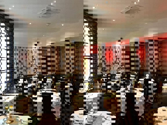 An interior view of the main dining room at Amber Regent. A large dining room with patterned, dark-red carpet and mainly cream walls. A monochrome botanical wallpapered wall and lattice work screens add dramtic accents to the room. The left wall also has a large, paned circular window. A dozen round tables, covered in white cloths and laid for dinner are scattered around the room, surrounded by dark upholstered, high-backed chairs.