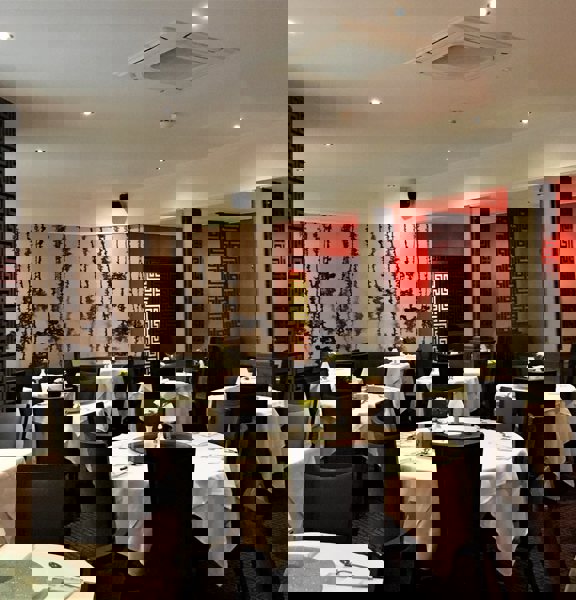 An interior view of the main dining room at Amber Regent. A large dining room with patterned, dark-red carpet and mainly cream walls. A monochrome botanical wallpapered wall and lattice work screens add dramtic accents to the room. The left wall also has a large, paned circular window. A dozen round tables, covered in white cloths and laid for dinner are scattered around the room, surrounded by dark upholstered, high-backed chairs.
