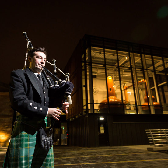 Piper playing in front of a distillery with copper kilns visible