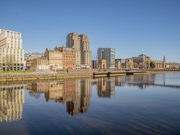 Sunny view across the River Clyde onto row of buildings, including the Clayton Hotel and the Virgin Hotel, with the buildings reflected in the river