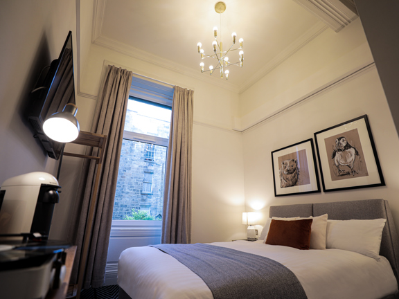 Compact hotel bedroom with double bed, high ceilings, graphic art work hands on the wall and modern coffee machine and record player sits on a wooden unit. 