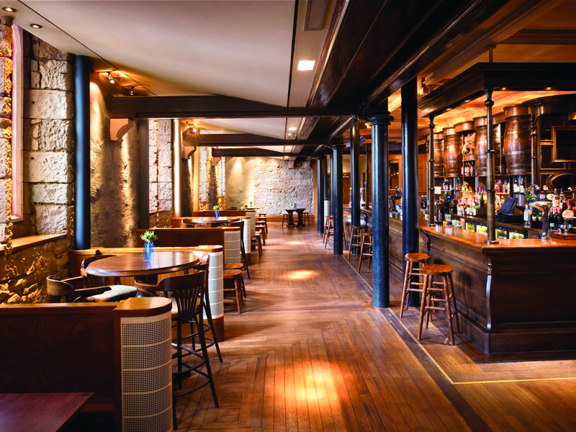 An interior of Oran Mor's bar space shows a room with bare stone walls and a low ceiling with wooden beams and black steel columns running the length of an old fashioned, wooden, panelled bar. The bar stretches the full length of the space, the fridges and shelves behind are fully stocked with bottles and 4 old fashioned wooden barrels are also visible on the top shelf. Tall wooden stools are placed intermittently along the bar, on the left tables with stools and chairs are placed beside each window.