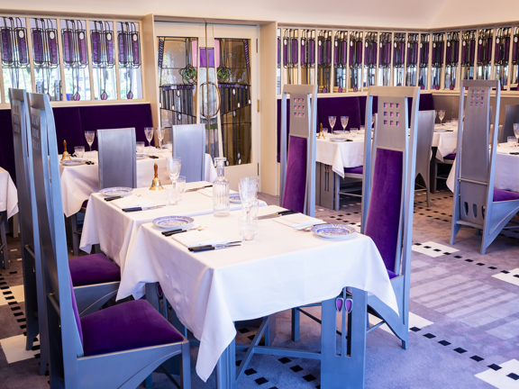 An internal view of The Salon De Luxe at Mackintosh at the Willow. The floor is carpeted with a luxurious lilac carpet, it has geometric, dotted patterns in shades of purple throughout. The top halves of the walls are lined with narrow stained-glass mirrors, with elongated, Glasgow-Style floral motifs. The double doors in the centre of the wall are, similarly, mirrored and embellished. Tables with white cloths are laid for a dinner; the chairs are incredibly tall and silver, with deep-purple velvet seats.