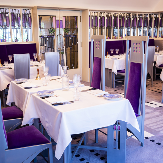 An internal view of The Salon De Luxe at Mackintosh at the Willow. The floor is carpeted with a luxurious lilac carpet, it has geometric, dotted patterns in shades of purple throughout. The top halves of the walls are lined with narrow stained-glass mirrors, with elongated, Glasgow-Style floral motifs. The double doors in the centre of the wall are, similarly, mirrored and embellished. Tables with white cloths are laid for a dinner; the chairs are incredibly tall and silver, with deep-purple velvet seats.