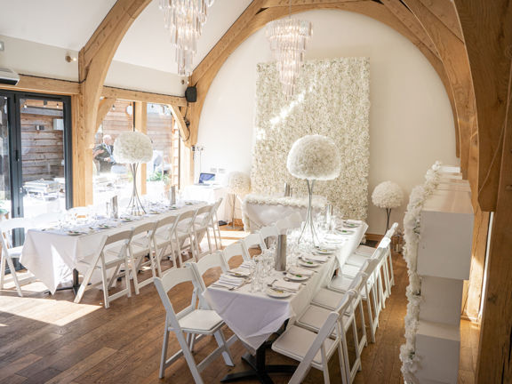 Barn venue space set with white tables and chairs and white flower wall arrangement. Wooden beams and chandeliers decorate the space.