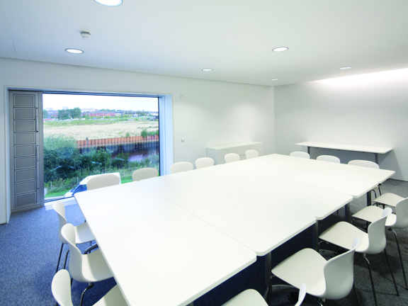An interior view of a private meeting space at the Riverside Museum. 4 white rectangular tables are arranged together, a dozen white plastic chairs with metal legs surround them. The room is carpeted with dark grey carpet and has white walls. The room is bright with spotlights in the ceiling and a large window in the long, far wall.