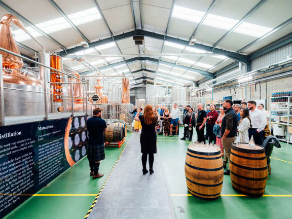 A group of people in a working distillery industrial pipe and containers as well as wooden whisky barrels can be seen. The group is being spoken to by a red haired guide. 