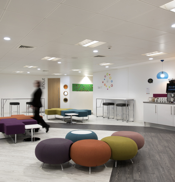 An photo of The Studio's break hall shows a large, bright interior space with white walls and wood effect flooring. Small islands of modern, brightly coloured seating in round and cross-shaped fabric pouffs are dotted throughout the space. The right hand wall has a long catering bar, with a coffee machine, mugs and tap. Glass blue and yellow ceiling lights hang above it. 2 high tables with stools are seen against the remaining wall space. A corridor and 2 doors can be seen off of the far wall of the space.