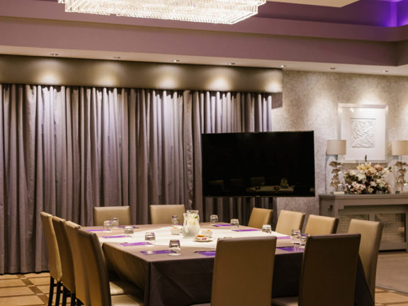 Conference room with square table and leather chairs around the table. Large TV screen in the corner. Large chandelier hangs above the table.  