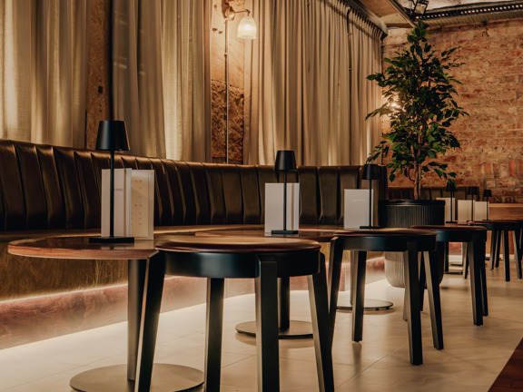 Room set up as a bar - exposed brick wall at one end and beige fabric hangs the length of the other wall. Low bar style table and chairs are dotted throughout the room.