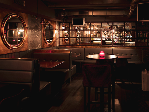 An interior from Blue Dog of booth seating, with high-backed leather upholstery around dark wooden tables. 2 large round mirrors with copper surrounds adorn the wall above each visible booth. The far wall is mirrored showing a reflection of a lit bar. Speakers and lights are mounted to the walls. 