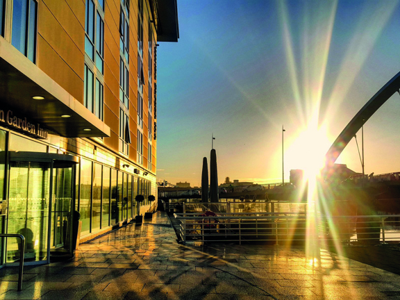 An early morning shot of the Hilton Garden Inn terrace shows the exterior of the hotel and the Clyde Arc bridge lit by the sun. The sun is low in the sky and its rays penetrate the image. The hotel ground floor is windowed floor to ceiling along the length of the visible wall. A glass revolving door with red lettering above, reading "Hilton Garden Inn" can be seen in the foreground. The paved floor is wet from rain and reflects the suns light. White railing run along the side of the River Clyde.