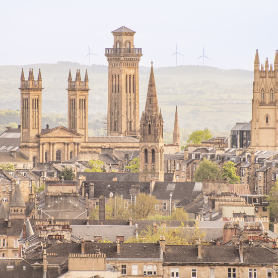 Cityscape of several church spires, including the four spires of the gothic Trinity Towers. 4 windmills can be seen on a hill in the background