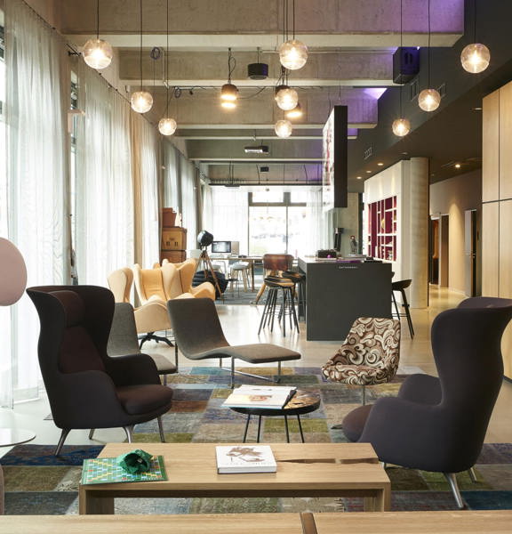 An interior view of the Moxy Merchant City lounge and reception area shows a bright, modern open plan space with glass, exterior walls. The floor is polished concrete, large rugs are placed in the seating areas. Eclectic, modern armchairs and coffee tables are dotted through the closer half of the space. A large TV can be seen mounted on a feature wall on the right. Beyond that a high counter painted grey is surrounded by high, dark metal stools; and beyond that more seating and shelves can be interpreted.