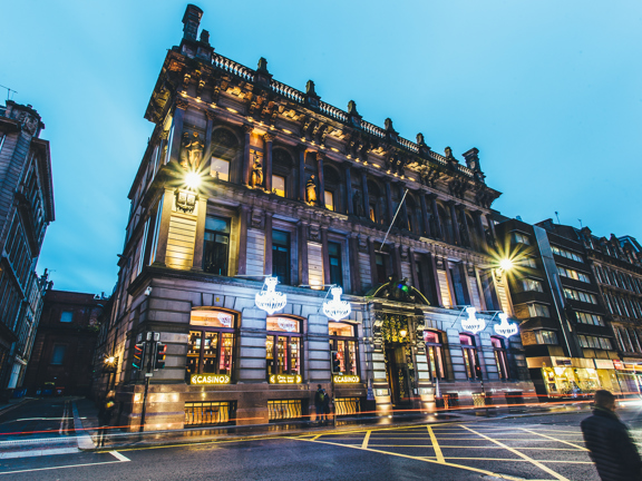 An exterior shot of Corinthian Rooms shows a large, stone building dominating a night street-scene. The building is up-lit, emphasising the carved figures and details on the buildings façade.  A few steps lead up to the main entrance, either side of which are 2 chandalier-style light fittings. Large windows adorn 3-storeys.