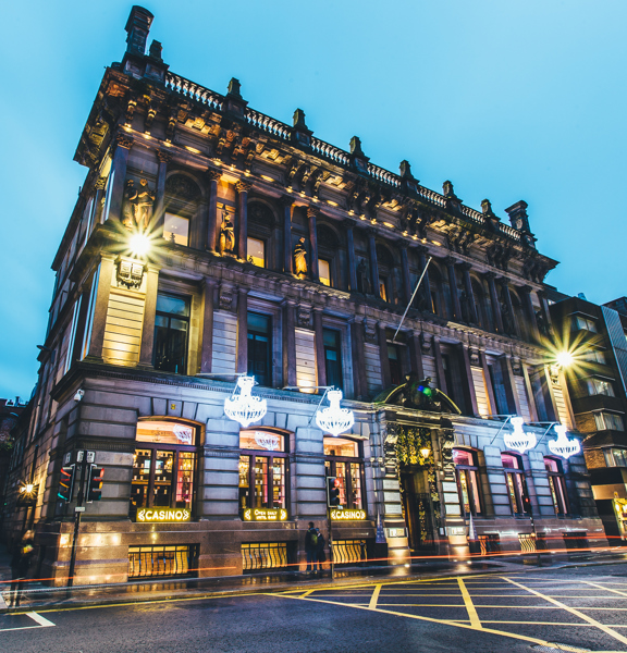 An exterior shot of Corinthian Rooms shows a large, stone building dominating a night street-scene. The building is up-lit, emphasising the carved figures and details on the buildings façade.  A few steps lead up to the main entrance, either side of which are 2 chandalier-style light fittings. Large windows adorn 3-storeys.