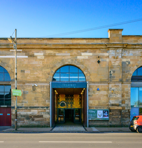 A photo of Tramway taken from over a road. A long, flat roofed Victorian, blonde sandstone building, with a huge central arched doorway and windows of the same scale on either side. The doorway has a grey sign reading "TRAMWAY" and set back from the street - above an automatic glass door - is a large yellow art work taking up the height of the entrance way. A parked car, bike racks, a lamppost and a CCTV camera can all be seen. The pavement is tarmac but the entrance to Tramway is paved with bricks.