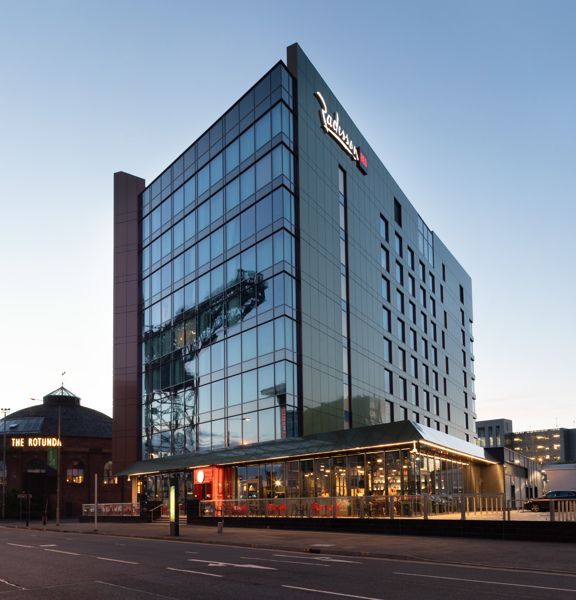 An exterior image of the Radisson Red hotel taken from over a road. A modern multi-storey building with a dark windowed front and dark grey, metallic cladding on the sides. A large lit-up sign of the Radisson Red logo adorns the top portion of this side wall. A narrow pavement runs the length of the road, off of which a few stairs lead to an entrance. A glass railing separates the pavement from a terrace with seating and a car park. At the end of the road buildings, including the SEC Armadillo are visible,
