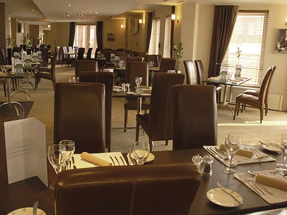 An internal view from Celtic Football Club shows a large, carpeted room decorated in neutral tones. There are dark tables and high-bcked chairs throughout the room with menus, napkins, silverware and glasses on the tables. Windows line one wall with plants, framed prints and a television screen also visible.