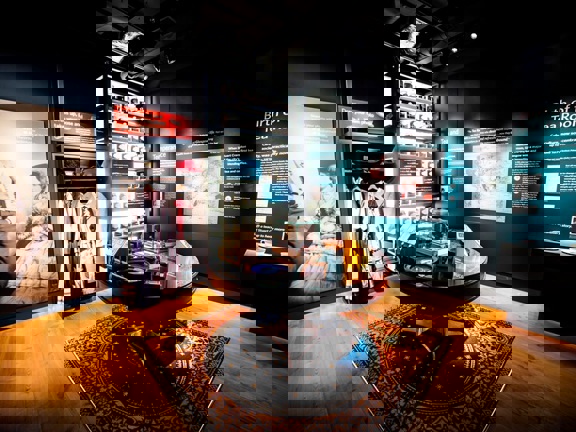 Dark walls with white writing tell the story of the tearooms. One wall has shelves of loose tea in jars that are light up from behind. Net to this is a range of traditional dress items from the 1900s. In the centre of the space is a wooden table with small wooden boxes of tea and interactive items.