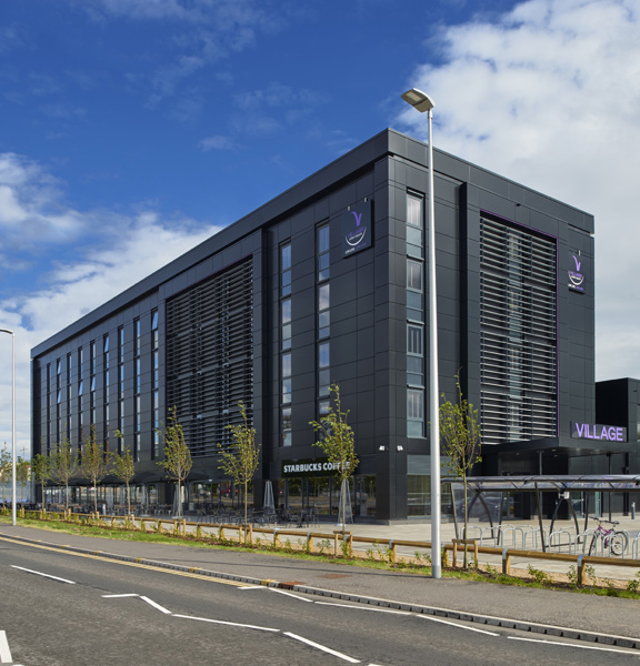 An image of the Village Hotel's exterior taken from across a road. Image shows a 4-storey, modern building with glass and black facing; against a blue, cloudy sky. A wide tarmac pavement runs along the either side of the curved road, a slim grassy verge, punctuated with saplings separates the pavement from the hotel carpark. A large covered bike rack is visible to the left of the entrance, recognisable by the purple "Village" branding above it. A Starbucks cafe is housed in the hotel's ground floor.