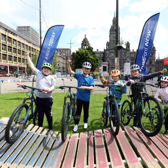 Five children stand next to their bikes cheering, in George Square, Glasgow.