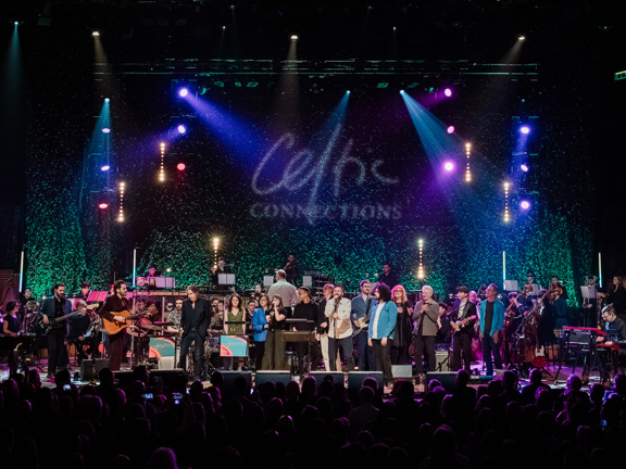 A large group of musicians perform on stage in front of Celtic Connections logo.