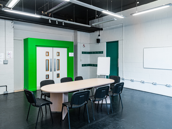 Room with black floor and white walls, a green frame surrounds the main entrance door. Black chairs surround an oval table a flip chart sits to the side.   