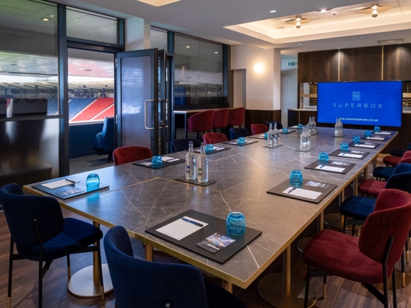 An interior view of a room at Hampden Stadium, a long marble effect table is surrounded by velvet red or blue chairs; stationery and glasses can be seen on the table. At the end of the table a large TV screen is on. The left hand wall of the room is made up of clouded glass. An open door onto a balcony allows us to see that the room is at the top of the stadium looking out at a view of the stadium seating.