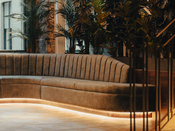 Long and winding brown suede sofa sits below floor to ceiling windows. Large green plants sit behind the sofa which is illuminated from below by a glow of light.
