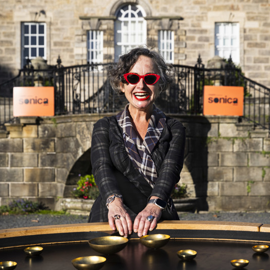 Cathie, Chief Executive of Cryptic dressed in tartan, smiling 4shows off an art installation outside Pollock House